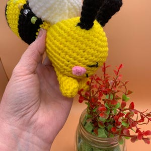 May include: A handmade crochet bee in yellow and black stripes, with white wings and black antennae. The bee has a pink mouth and green eyes. The bee is being held in a hand. The background is a solid peach color.