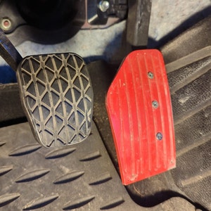 Puede incluir: Primer plano de dos pedales de coche. El pedal izquierdo es negro con un patrón de diamante y el pedal derecho es rojo con una superficie acanalada.