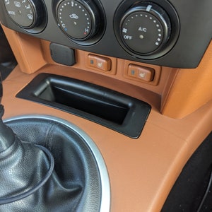 Può includere: Primo piano dell'interno di un'auto con un cruscotto in pelle marrone, un cambio marcia nero e un pannello di controllo nero con pulsanti per il controllo del clima e i sedili riscaldati. Il testo "A/C" e "OFF" è visibile sul pannello di controllo.