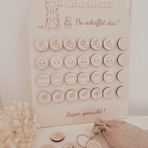 Könnte beinhalten: Eine hölzerne Belohnungstafel für das Töpfchentraining mit dem Namen "Charlotte" und dem Satz "Du schaffst das!". Die Tafel zeigt gravierte Symbole wie Tropfen, Kack-Emojis, Toilettenpapier, Waschbecken und Herzen. Ein kleiner Jutebeutel und zusätzliche Token sind ebenfalls vorhanden.