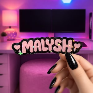 May include: A pink and black name tag with the name "MALYSH" in a bubble font. The name tag has a black border with glitter and heart and star accents. The name tag is held in front of a desk with a computer monitor.