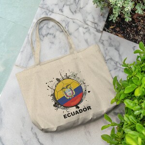 May include: A beige tote bag featuring a black and white graphic of a football with the Ecuadorian flag on it. The text "ECUADOR" is printed below the graphic.