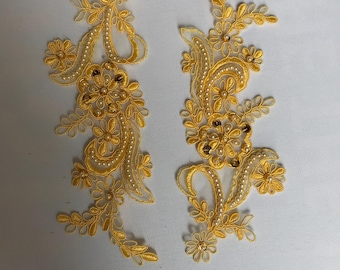May include: Two golden-yellow floral lace appliques. Each applique features a detailed design of flowers, leaves, and swirling patterns, embellished with small white beads and gold sequins. The appliques are set against a light background.