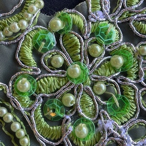 May include: Close-up of a green and silver embroidered fabric with pearl beads and sequins. The fabric has a floral design with a central flower.