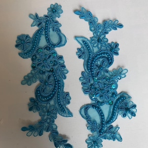 May include: Two turquoise lace appliques with floral and paisley designs. Each applique features intricate embroidery, sequins, and small bead accents. The appliques are set against a white background, highlighting their detailed craftsmanship.