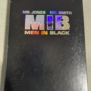 May include: Black DVD case for the movie "Men in Black." The title is in large, iridescent letters with the acronym "MIB." The DVD case also features the names "Mr. Jones" and "Mr. Smith."