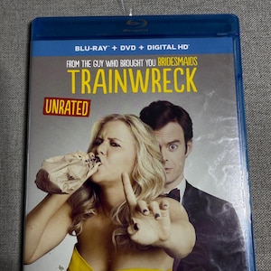 May include: A blue Blu-ray case for the movie "Trainwreck" featuring Amy Schumer and Bill Hader. The cover art shows Schumer with a paper bag over her mouth and Hader in a suit. The case includes "Blu-ray + DVD + Digital HD".