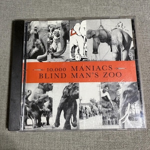 May include: A CD case featuring the album "Blind Man's Zoo" by 10,000 Maniacs. The cover art is a collage of black and white images of elephants, with the album title and band name in white text on a red banner.