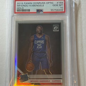 May include: A 2019 Panini Donruss Optic basketball card featuring Mfiondu Kabengele of the Clippers. The card is graded GEM MT 10 and encased in a protective holder. The card has a holographic design and the text "RATED ROOKIE".