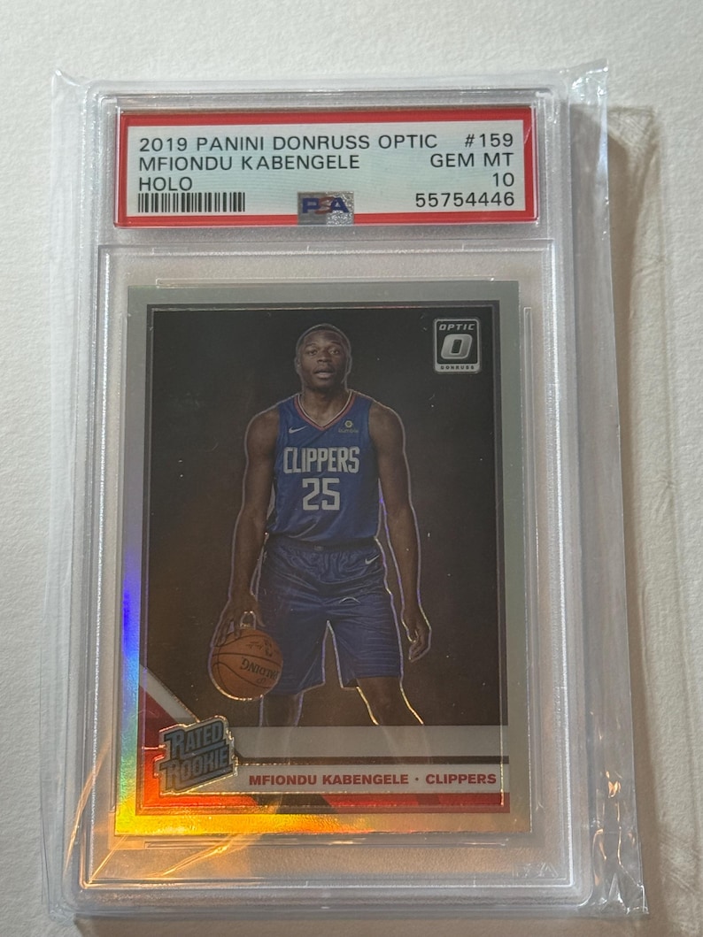 May include: A 2019 Panini Donruss Optic basketball card featuring Mfiondu Kabengele of the Clippers. The card is graded GEM MT 10 and encased in a protective holder. The card has a holographic design and the text "RATED ROOKIE".