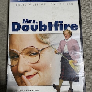 May include: DVD case for the movie "Mrs. Doubtfire" featuring Robin Williams and Sally Field. The cover shows a close-up of a woman wearing glasses and a full-body image of Mrs. Doubtfire holding a broom. Text includes "BEHIND-THE-SEAMS EDITION" and "SHE'LL ROCK YOUR WORLD!"