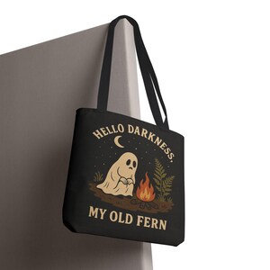 May include: Black tote bag featuring a cartoon ghost sitting by a campfire under a crescent moon. The text "HELLO DARKNESS, MY OLD FERN" is printed above and below the illustration. The bag has black handles.
