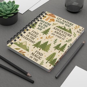 May include: A cream-coloured spiral-bound notebook with a nature-themed design. The cover features illustrations of trees, mountains, mushrooms, and text that reads "Breathe in the wild air" and "Adventure awaits, go find it."