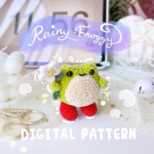 Könnte beinhalten: Ein Häkelmuster für einen grünen Frosch mit roten Stiefeln, der eine weiße Blume hält. Das Muster heißt "Rainy Froggy" und ist ein digitaler Download.