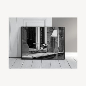 May include: Black and white photograph of a window with a lamp hanging above it. The window is framed in stone and has a wooden sill. There are flowers on the sill and a small wooden object in the window.