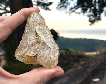 May include: A clear, translucent, irregularly shaped crystal with a rough, textured surface. The crystal is held in a hand against a blurred background of trees and a distant landscape.