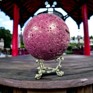 May include: A large, purple, sphere-shaped, decorative stone with a rough, textured surface. The stone is on a silver metal stand with ornate details.
