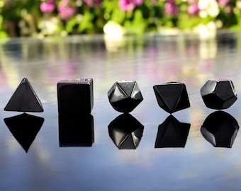 May include: A collection of five black geometric shapes, including a pyramid, cube, and faceted forms, are arranged in a row. The objects are highly reflective, with their mirrored images visible below. The background features a blurred view of colorful flowers.