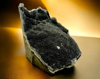 May include: A dark, textured mineral specimen with a rough, granular surface. The stone is primarily black with areas of lighter gray and white. The mineral has a jagged, irregular shape and sits on a gold-toned surface.