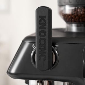 May include: Close-up of a black espresso machine with a coffee bean grinder. The machine features a black handle with the word "KNOCK" engraved on it. The machine has a sleek, modern design.