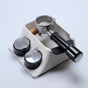 May include: A white coffee station organizer with a portafilter, two tampers, and a holder. The portafilter is stainless steel with a black handle. The tampers are silver and black. The organizer is on a white surface.