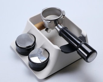 May include: A white coffee station organizer with a portafilter, two tampers, and a holder. The portafilter is stainless steel with a black handle. The tampers are silver and black. The organizer is on a white surface.