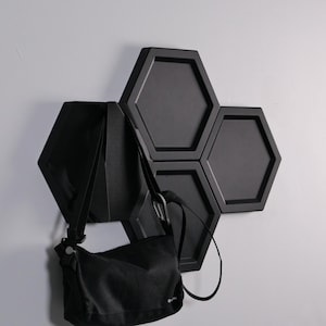 May include: A black, hexagonal wall-mounted coat rack. A black shoulder bag hangs from the rack. The rack is made of a matte black material and is mounted on a white wall. The design is geometric.