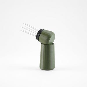 May include: A green coffee distribution tool with a black ring and thin metal needles. The tool is composed of two parts, a cylindrical base and a top section angled to the side. The tool is designed to break up clumps in ground coffee.