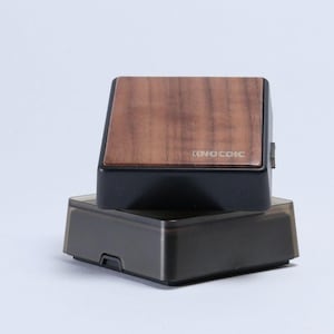 以下が含まれることがあります： 前面に「KNOCDIC」の文字が入った黒と茶色の長方形のデバイスで、透明な長方形のベースの上に置かれています。デバイスの上部は木目調です。ベースは透明でスモーキーな色です。
