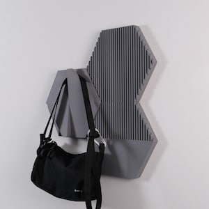 May include: A gray, geometric wall-mounted coat rack with a black bag hanging from it. The rack has a hexagonal shape with vertical lines on one section. The bag has a black strap and a silver-colored clasp.