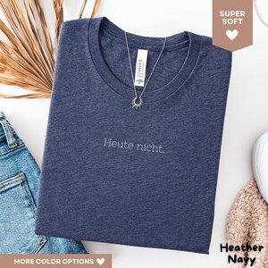 May include: Heather navy blue t-shirt with the phrase "Heute nicht" in white script. A silver sunburst pendant necklace rests on the shirt. The shirt is folded, with a pair of blue jeans visible.