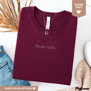 May include: A maroon t-shirt with the text "Heute nicht." in white. A silver necklace with a sunburst pendant is draped over the shirt. The shirt is folded and displayed on a white surface, with a pair of blue jeans and a beige sweater visible.