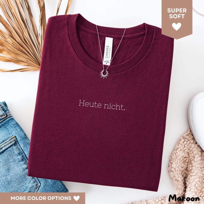 May include: A maroon t-shirt with the text "Heute nicht." in white. A silver necklace with a sunburst pendant is draped over the shirt. The shirt is folded and displayed on a white surface, with a pair of blue jeans and a beige sweater visible.