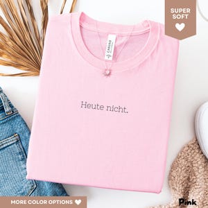 May include: A light pink t-shirt with the German phrase "Heute nicht" printed in black. The shirt has a crew neck and is made of soft material. A silver necklace with a sun pendant is visible.