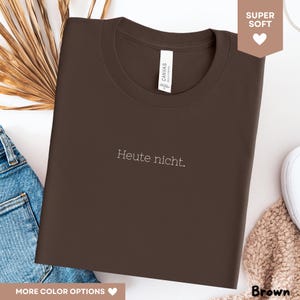 May include: A brown t-shirt with the text "Heute nicht" in white. The shirt has a crew neck and a tag that says "Canvas". The image also shows a pair of blue jeans, a white shoe, and a brown knitted item.