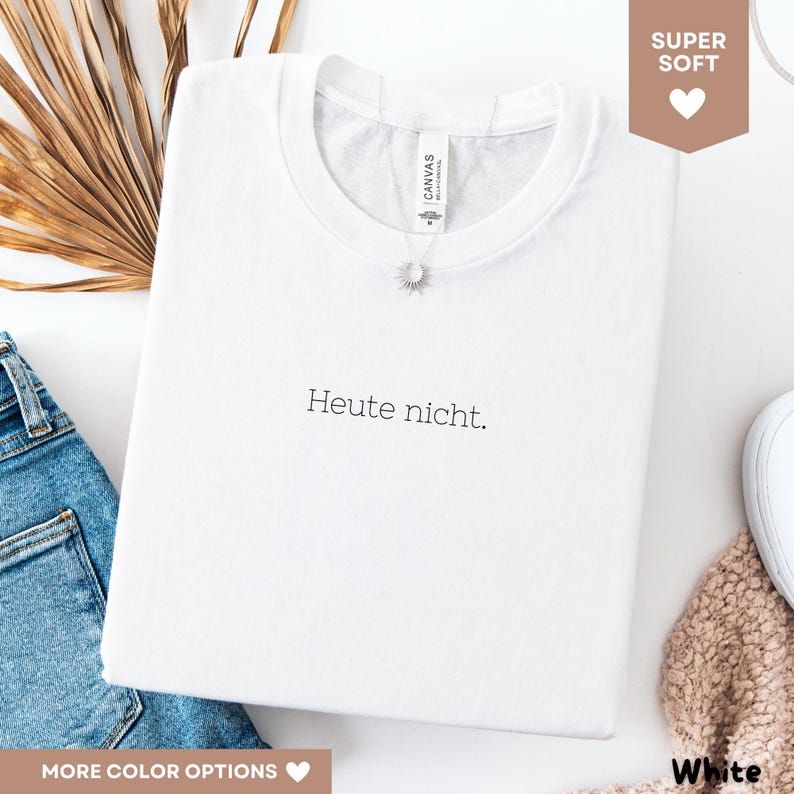 May include: A white crew neck t-shirt with the German phrase "Heute nicht" printed in black. The shirt is folded and has a small silver pendant necklace. The image also shows a pair of blue jeans and a white shoe.