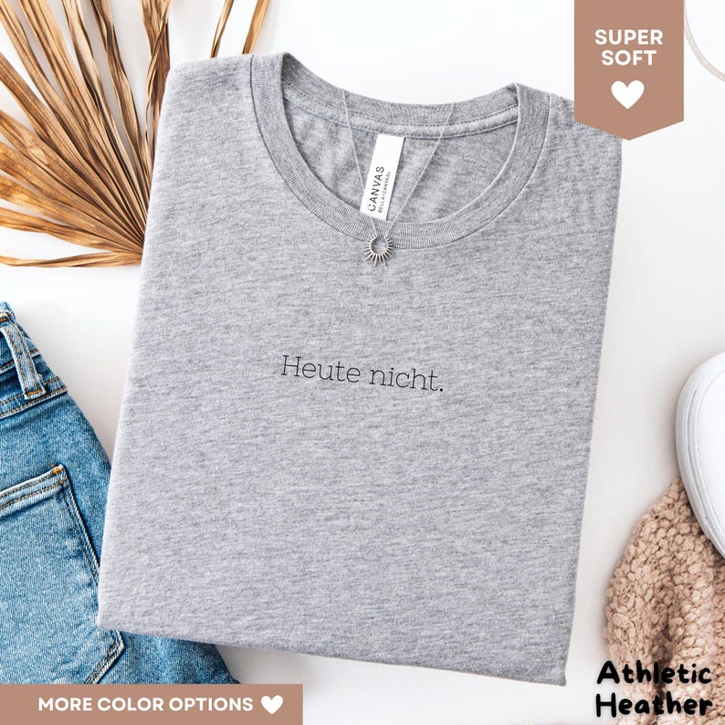 May include: A heather gray t-shirt with the German phrase "Heute nicht" printed in black. The shirt has a crew neck and is paired with a silver necklace. The words "Super Soft" and "Athletic Heather" are also visible.