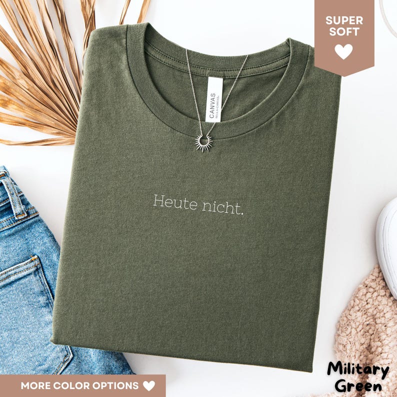 May include: Military green t-shirt with the embroidered phrase "Heute nicht." A silver necklace with a sun pendant is draped over the shirt. The shirt is folded and placed on a white surface.