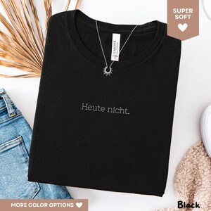 May include: Black t-shirt with the phrase "Heute nicht" in white script. A silver necklace with a sunburst pendant is draped over the shirt. The shirt is folded, and a pair of blue jeans is visible in the background.