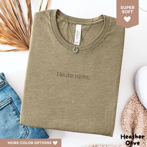May include: Heather olive-colored t-shirt with the German phrase "Heute nicht" printed in black. A silver necklace with a sun pendant hangs on the shirt. The shirt is folded and displayed with a pair of blue jeans and a white shoe.