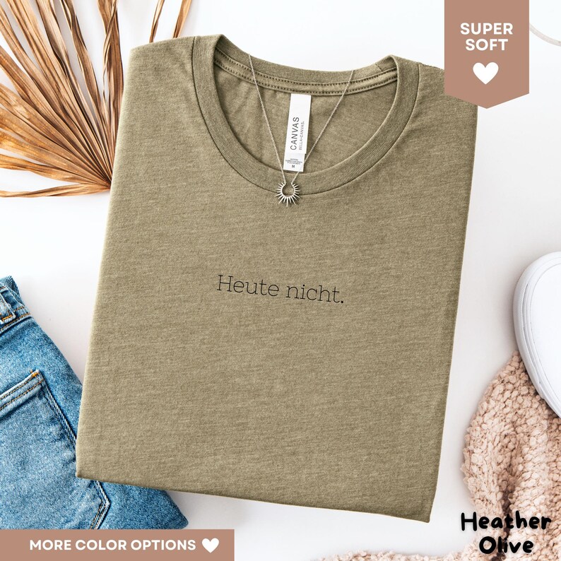 May include: Heather olive-colored t-shirt with the German phrase "Heute nicht" printed in black. A silver necklace with a sun pendant hangs on the shirt. The shirt is folded and displayed with a pair of blue jeans and a white shoe.