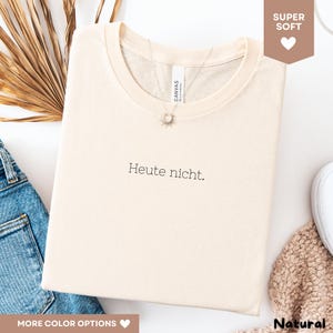 May include: Cream-colored t-shirt with the German phrase "Heute nicht" printed in black. The shirt is folded and has a round neckline. A silver necklace with a sun pendant is visible. The shirt is made of a soft material.