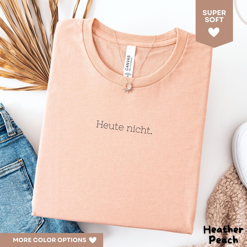 May include: A peach-colored t-shirt with the text "Heute nicht." in black. The shirt has a crew neck and is made of soft material. A silver necklace with a sun pendant is visible. The shirt is folded on a white surface.