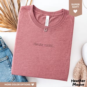 May include: Heather mauve t-shirt with the text "Heute nicht." A silver necklace with a sunburst pendant is draped over the shirt. The shirt is folded and placed on a white surface. The shirt is made of a soft material.