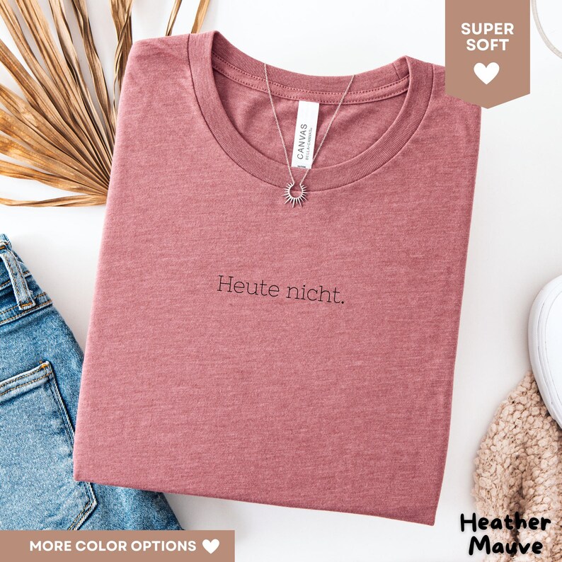 May include: Heather mauve t-shirt with the text "Heute nicht." A silver necklace with a sunburst pendant is draped over the shirt. The shirt is folded and placed on a white surface. The shirt is made of a soft material.