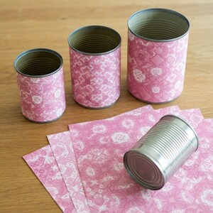 May include: Three cylindrical metal cans of varying heights wrapped in pink paper with a white geometric floral pattern, and one additional plain silver metal can lying on its side on top of several sheets of the same pink patterned paper.