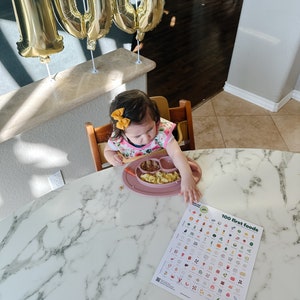 May include: A young child sits in a high chair at a marble table, eating from a pink plate.  The child is wearing a pink floral bib and a yellow bow in their hair.  A chart titled "100 first foods" is on the table.  There are gold balloons in the background that spell out "100".