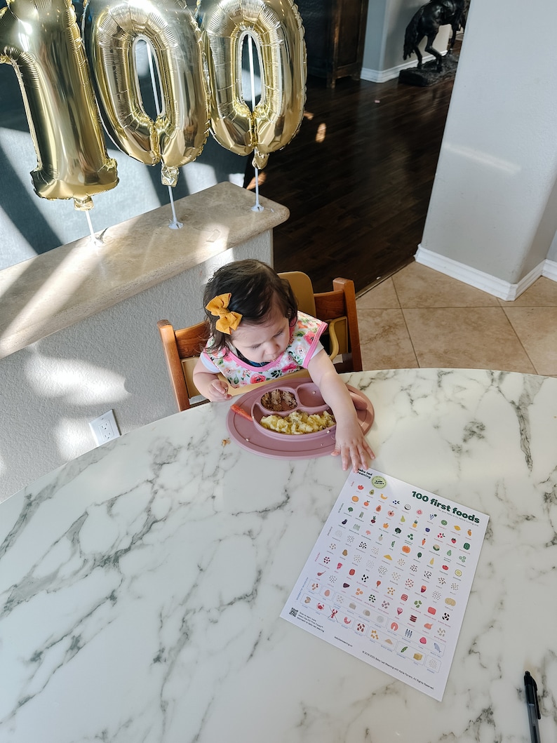 May include: A young child sits in a high chair at a marble table, eating from a pink plate.  The child is wearing a pink floral bib and a yellow bow in their hair.  A chart titled "100 first foods" is on the table.  There are gold balloons in the background that spell out "100".