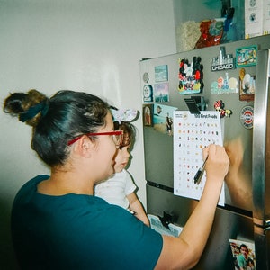 May include: A woman and a young child are standing in front of a refrigerator. The woman is pointing at a chart on the refrigerator that says "100 First Foods". The chart has pictures of different foods on it.
