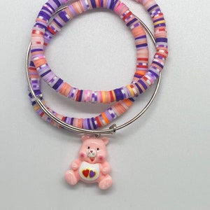 May include: A set of two colourful beaded bracelets with a pink bear charm. The bracelets are made of small, round beads in shades of purple, pink, orange, and white. The bear charm has a purple heart on its chest.